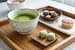 Bol en céramique contenant du matcha vert mousseux, posé sur un plateau en bois avec des wagashi japonais, dont des mochis verts et des sucreries traditionnelles, dans un cadre harmonieux de dégustation du thé